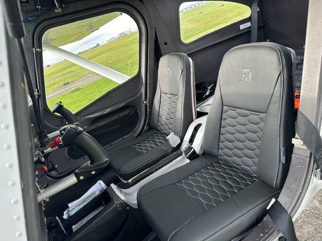 Black quilted aviation cockpit seats installed inside a Cessna C162 cabin, with the pilot's seat in front and a window view of green grass outside.