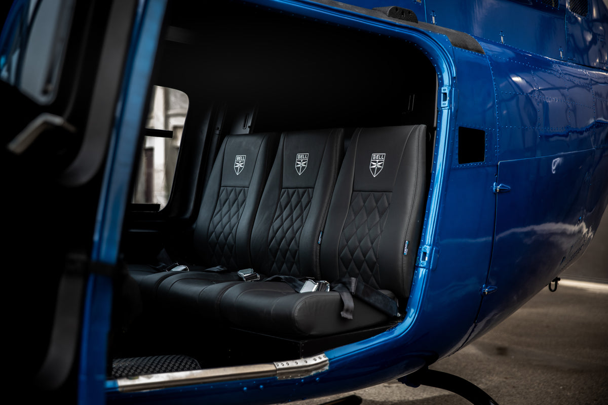 Close-up of a blue helicopter interior with a black quilted three-seat bench and embroidered crest logos on the backrests, shown with the door open.